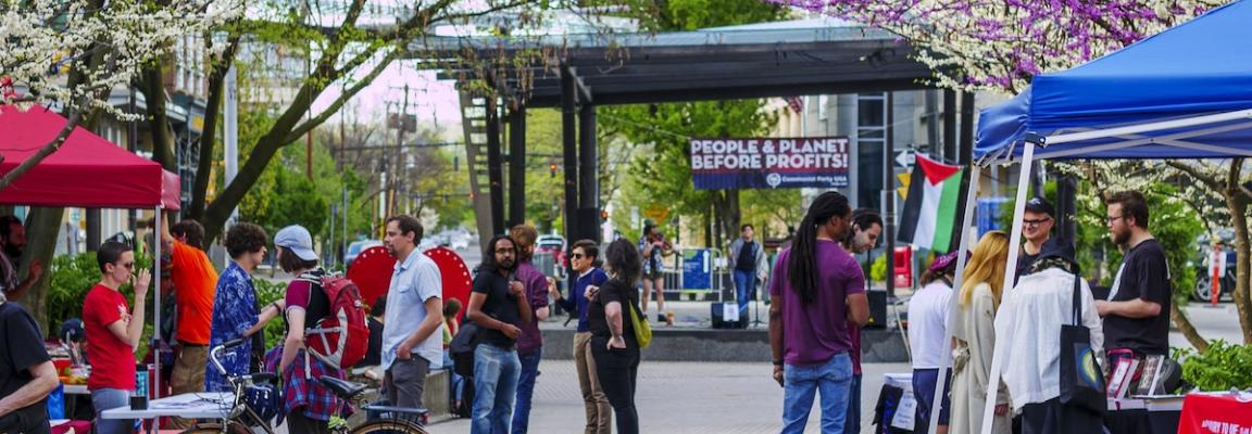 Ithaca progressives celebrate Mayday 2024 at Bernie Milton Pavillion on the Ithaca Commons. A crowd of attendees mingle with organizers at their tables in front of the pavillion stage. Trees/flowers are in full bloom with Ithaca CPUSA banner in the background